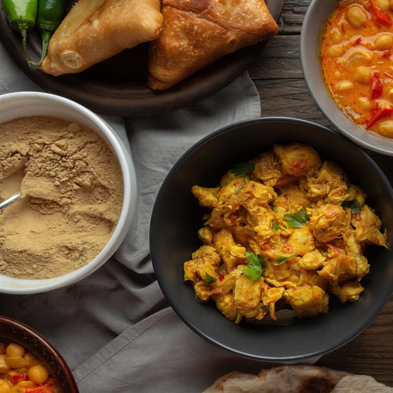 Overhead view of Indian dishes: chicken curry, chickpea curry, samosas, and spices on a wooden table.