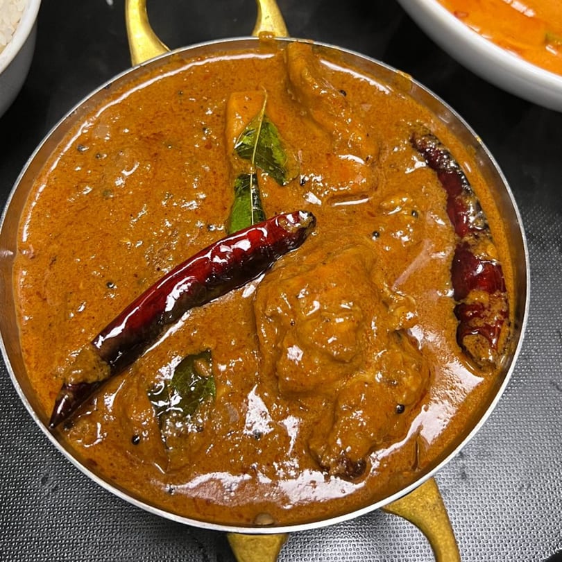 Close-up of a rich, orange-brown curry with whole red chilies and green leaves in a metal bowl.