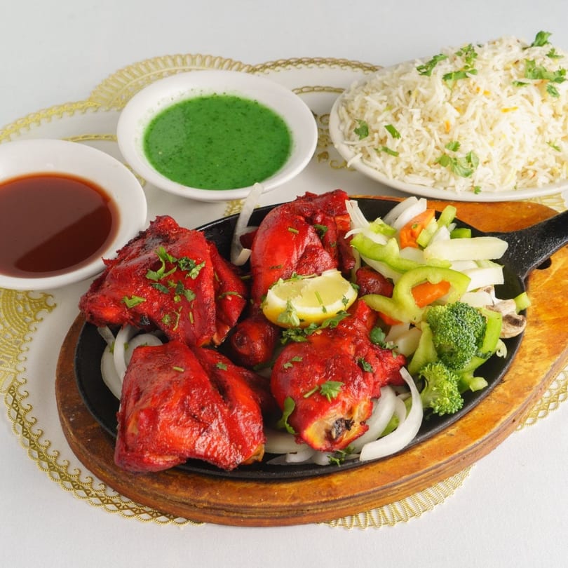 Red chicken tikka with vegetables, rice, and two dipping sauces on a white tablecloth.
