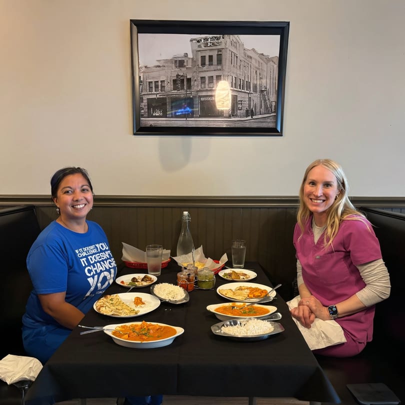 Two women smiling at a table laden with Indian food, including curries, rice, and naan.