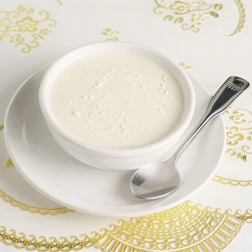 A white bowl of creamy yogurt with a spoon, on a white plate with a gold patterned placemat.