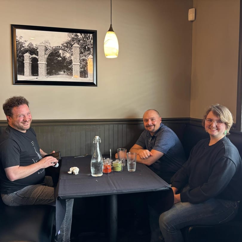 Three smiling people sit at a restaurant table with a black tablecloth, water, and spices.