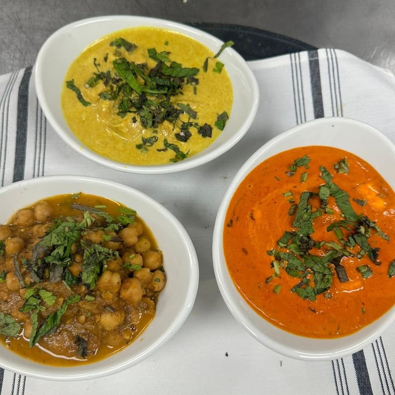 Three bowls of Indian food, including yellow curry, chickpea curry, and red curry, garnished with herbs.